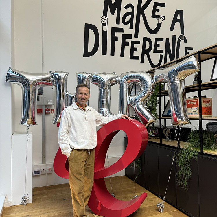 MD and Founder Ollie Leggett standing in front of a giant 3D red IE logo. Large silver balloons behind him spell THIRTie. Above him on the wall of the studio is a mural saying "Make a Difference" 