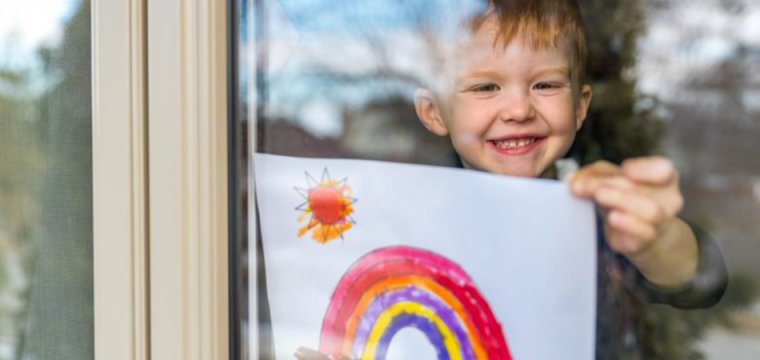 Photo of a little boy sticking a drawing of a rainbow of a rainbow in the window of his house, something that became popular during the Covid-19 lockdowns