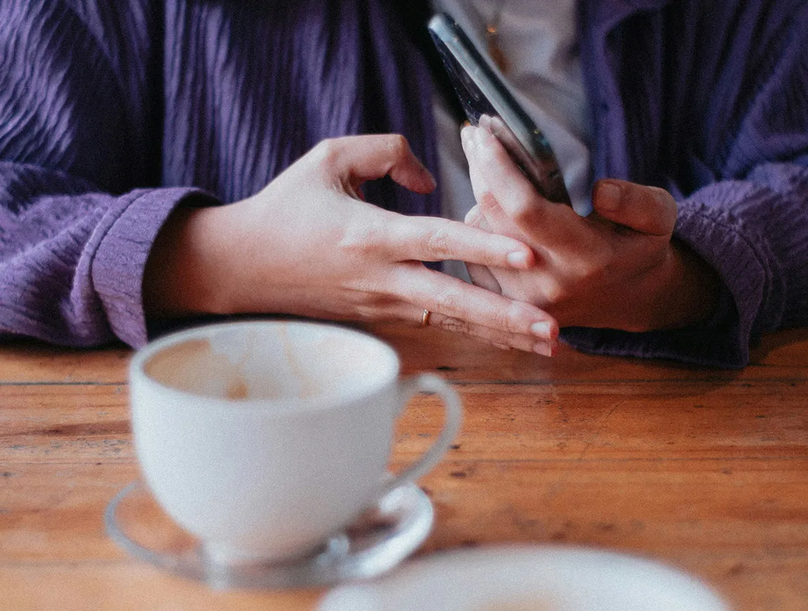 Close up on someone's hands holding a mobile phone, with a coffee cup on the table in front of them